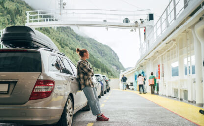 Woman leans against her car on Norwegian ferry surrounded by other vehicles with misty green mountains in background. Scene captures essence of relaxed travel, exploration in scenic Norwegian fjords